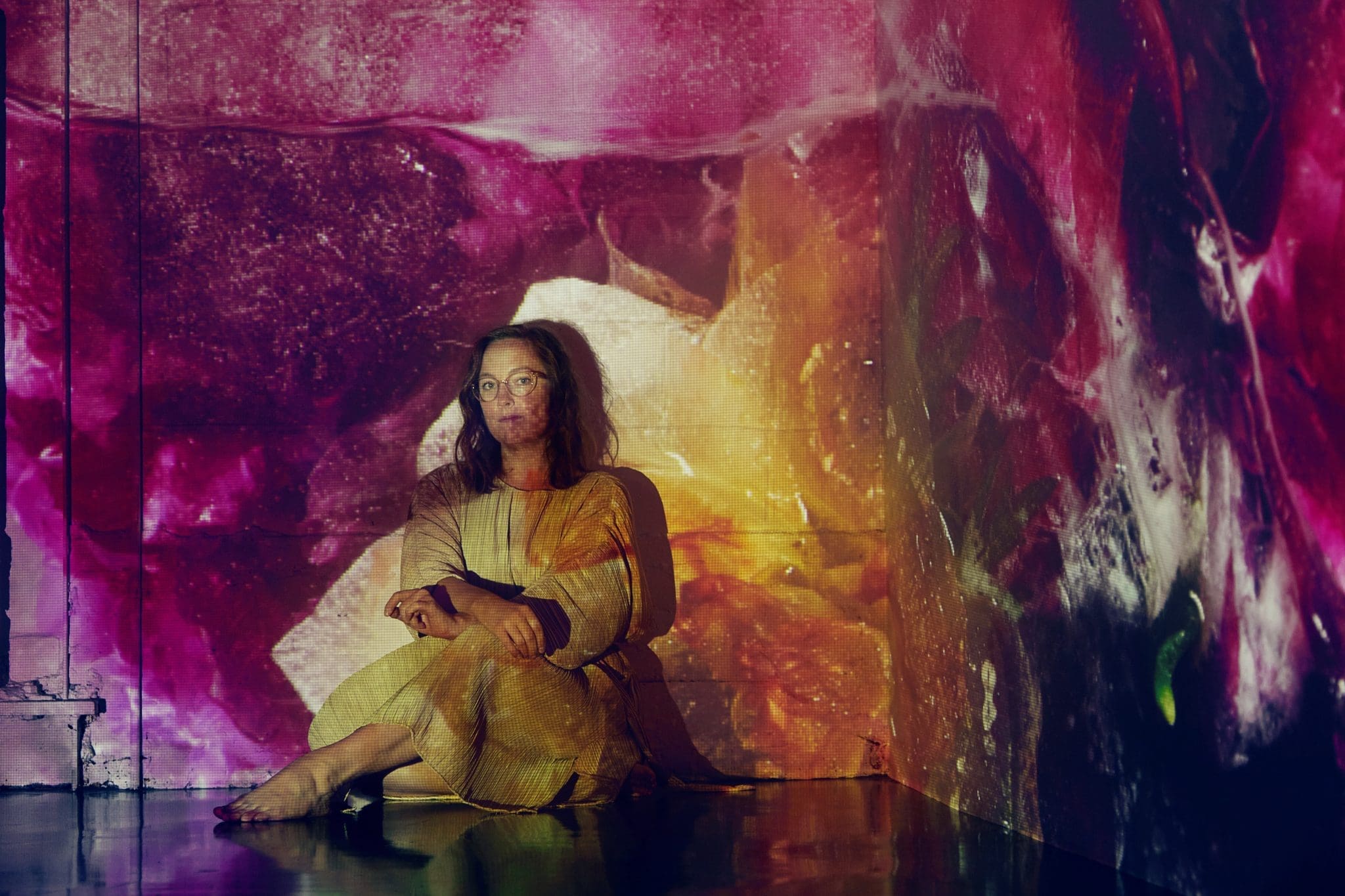 Press photo of Sarah Hiltz sitting on the floor with a projection of a fire and flowers over her